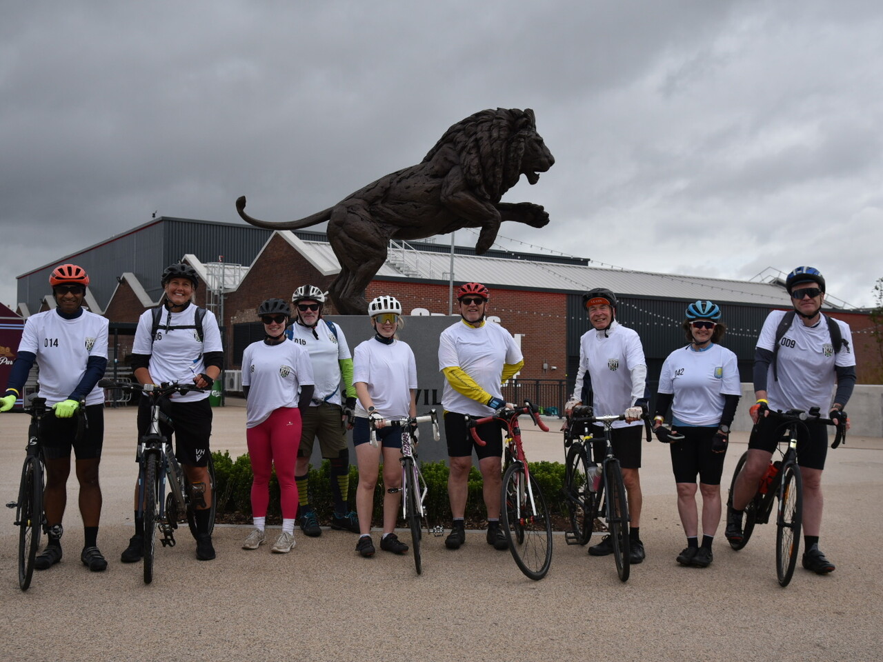 Nine cyclists stand with their bikes in front Villa Park posing for a group photo.