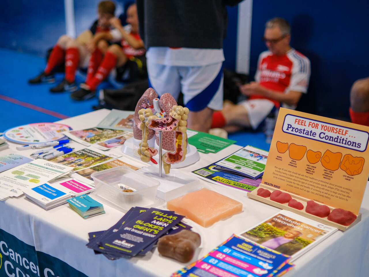 Let's Talk Hope table displaying prostate cancer awareness materials at an indoor sports venue, including anatomical models, informational leaflets.