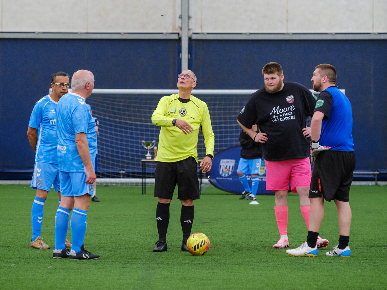 Referee in a bright yellow shirt stands on an outdoor pitch with a football at their feet, speaking to two small groups of players in blue and dark kits gathered for a pre-match coin toss, with a goal and trophy table visible behind.