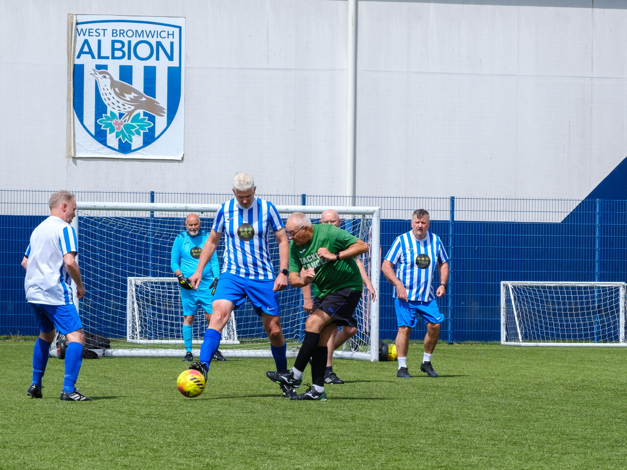 Wide shot of Big C tackling Cancer and Movers and Shakers players on the pitch with WBA logo on the dome in the background.