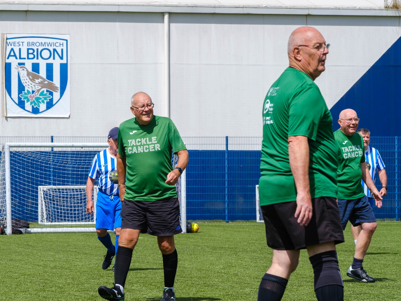 Close-up of Big C tackling Cancer players on the pitch with WBA logo on the dome in the background.