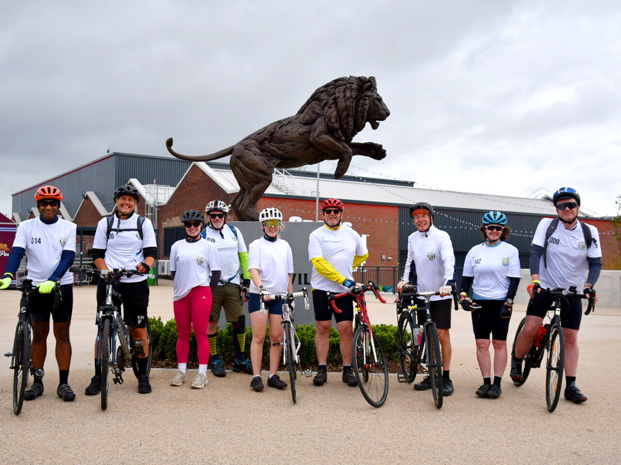 Nine cyclists stand with their bikes in front Villa Park posing for a group photo.