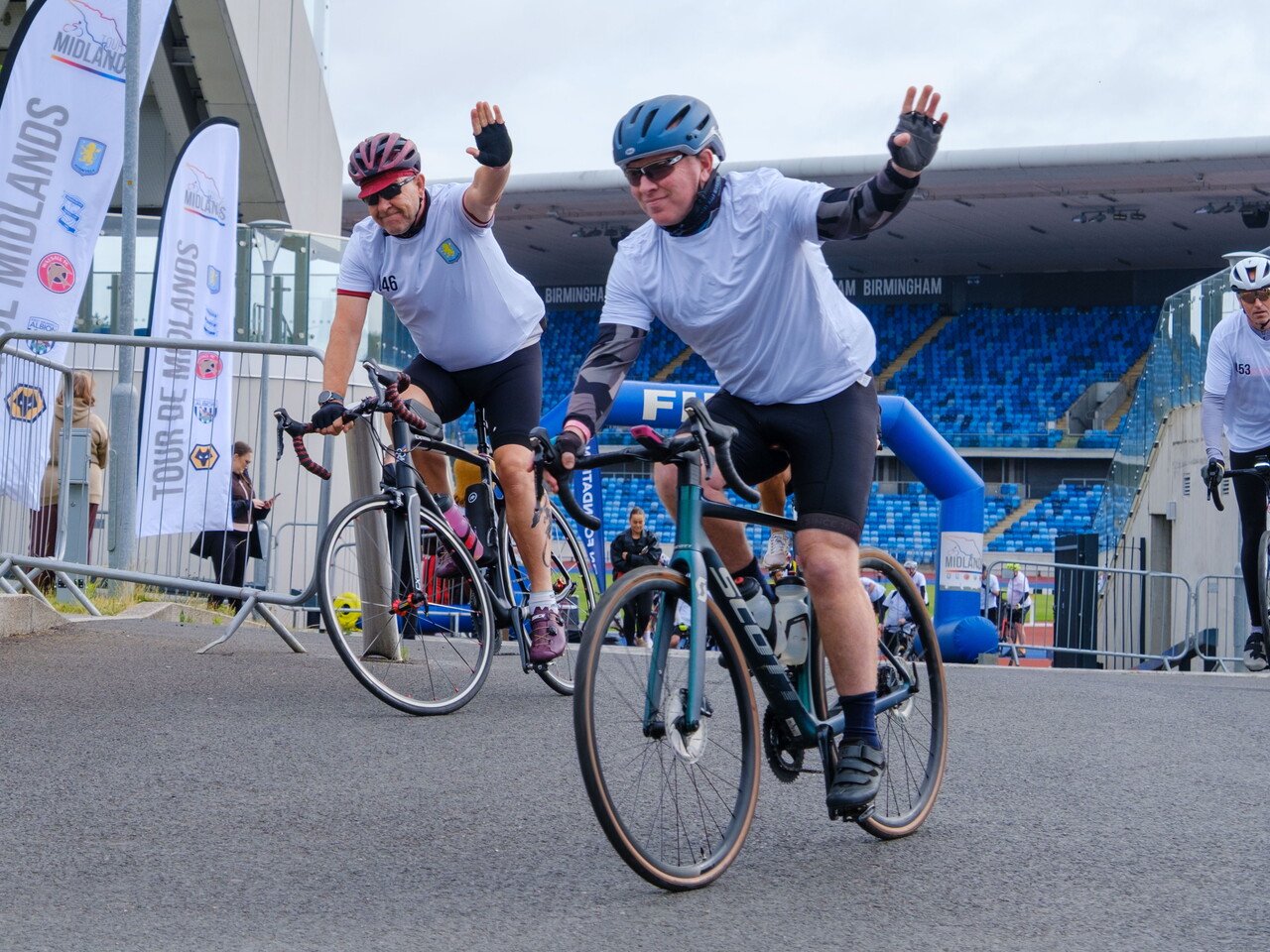  Two cyclists wave to the camera while while cycling out of Alexander Stadium