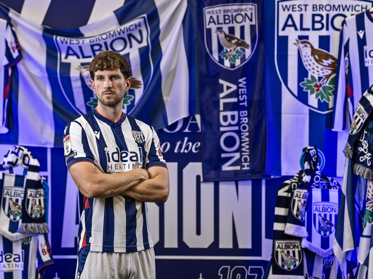 Chris Mepham in the home kit looking at the camera with his arms folded while stood in front of an Albion back drop with plenty of WBA branding