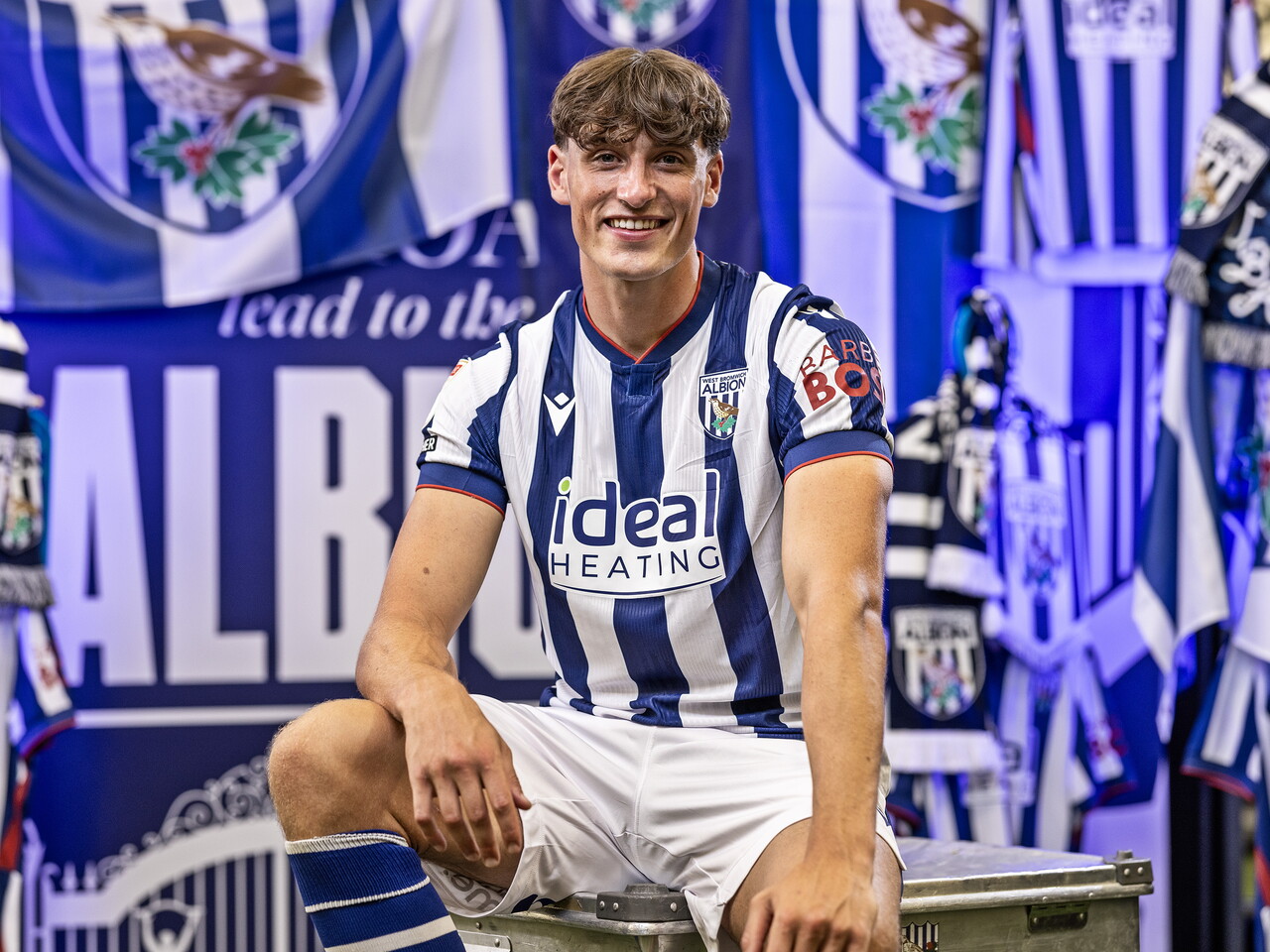 Barney Stewart in the home kit smiling at the camera while sat in front of an Albion back drop with plenty of WBA branding