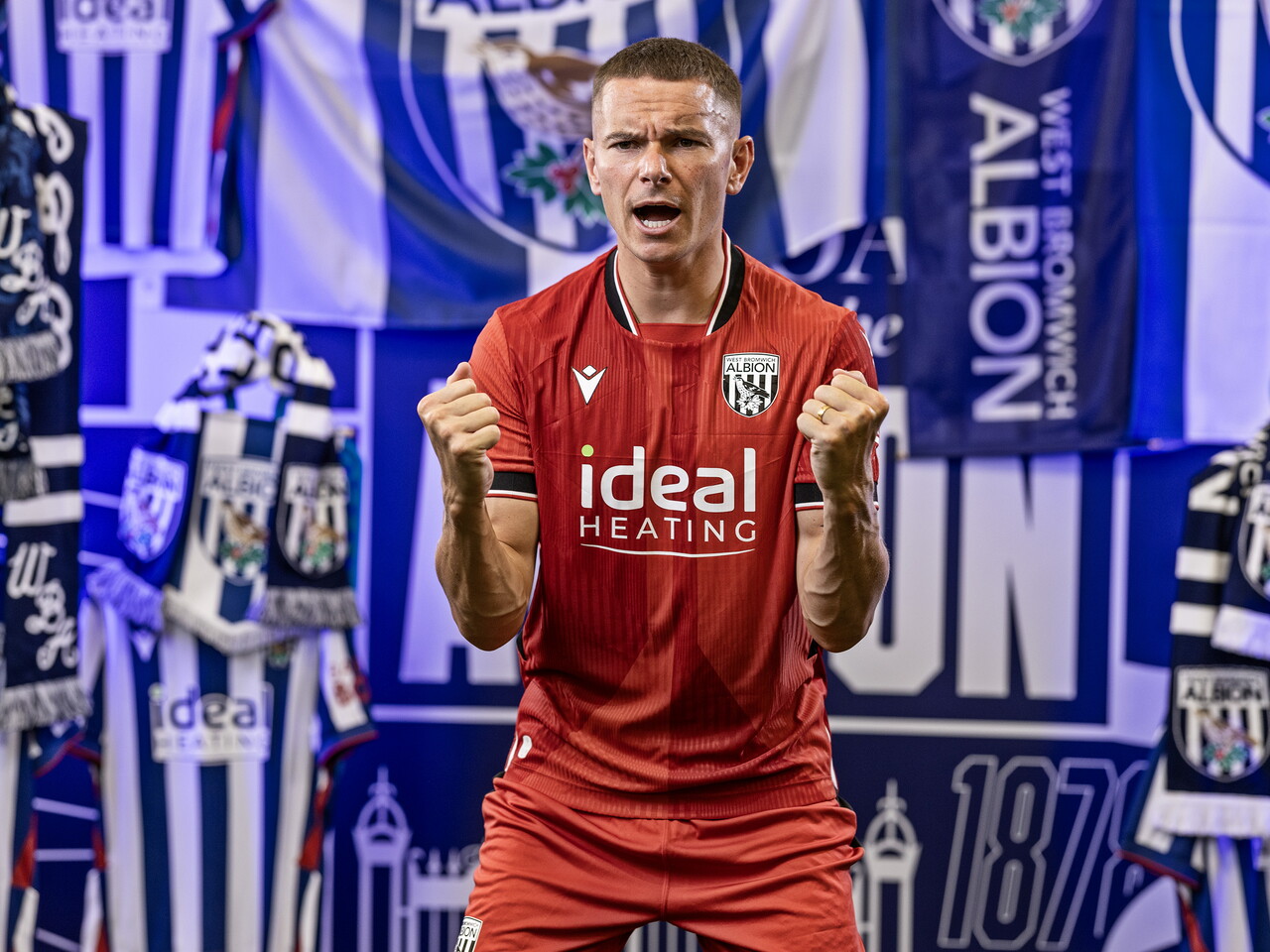 Conor Townsend in the red away kit celebrating towards the camera while stood in front of an Albion back drop with plenty of WBA branding