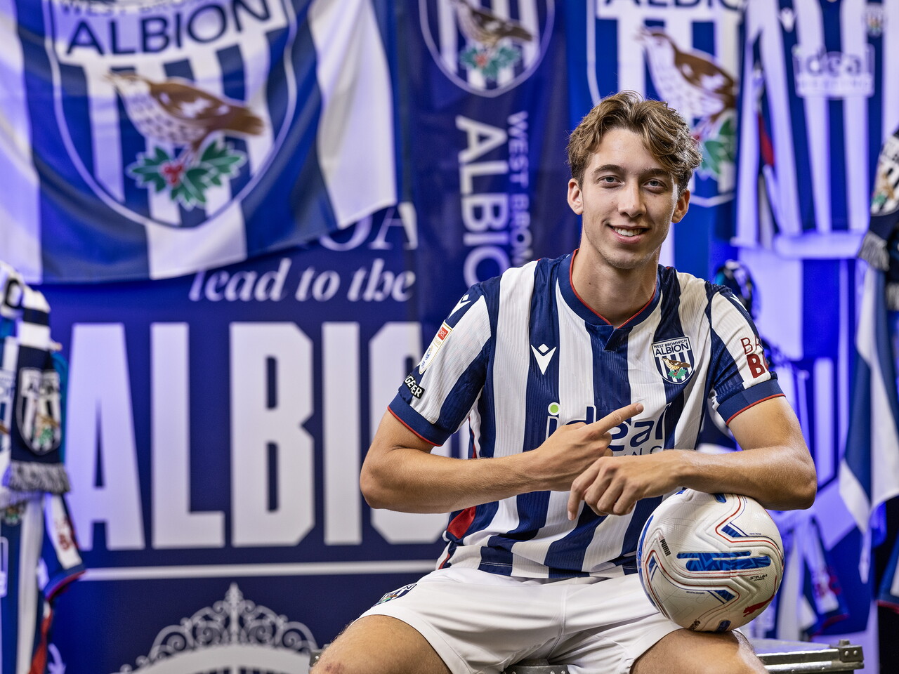 Harry Whitwell in the home kit smiling at the camera and leaning on a ball while sat in front of an Albion back drop with plenty of WBA branding