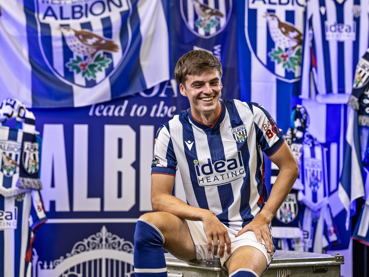 Alex Williams in the home kit laughing while sat in front of an Albion back drop with plenty of WBA branding