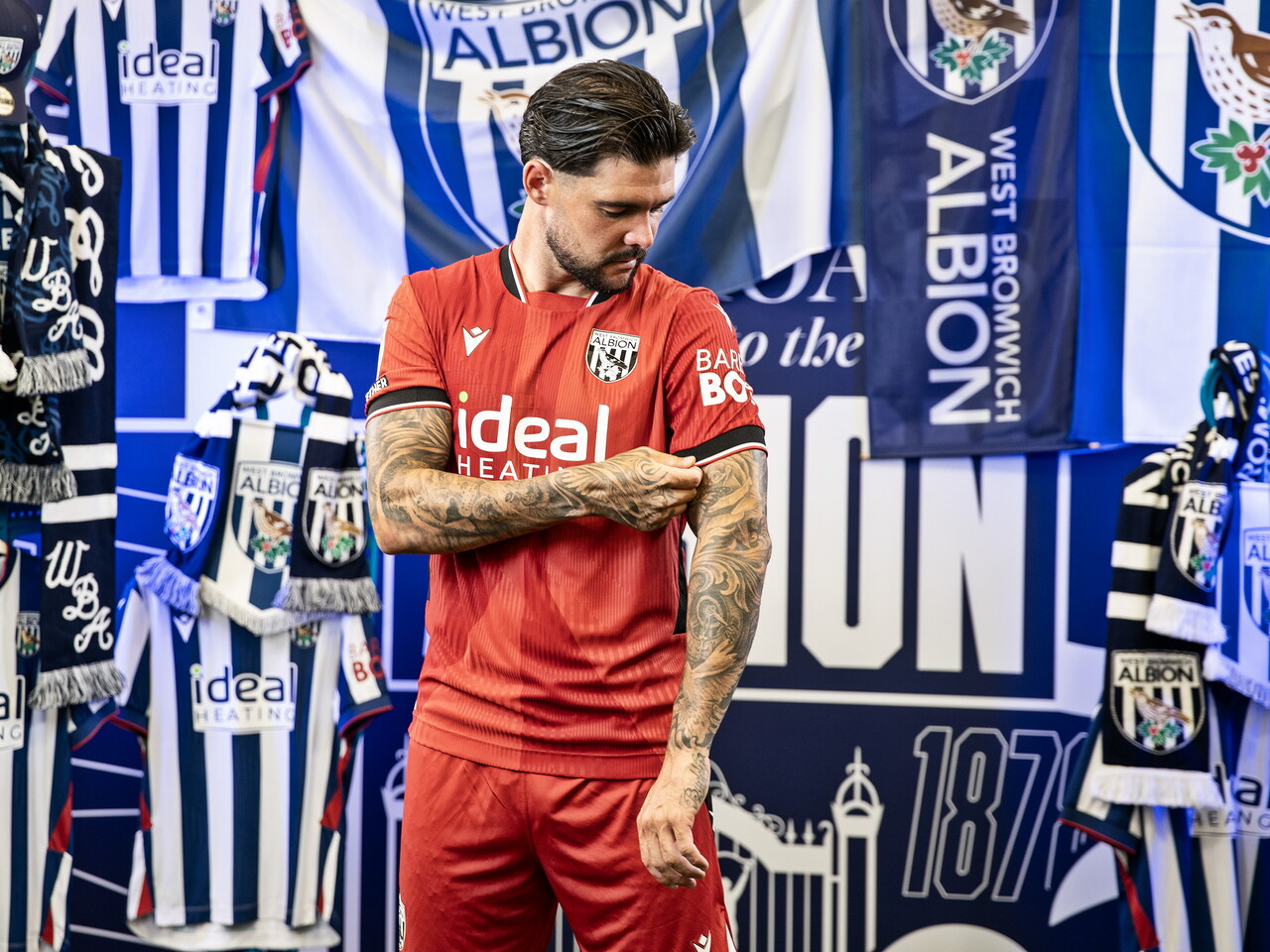Alex Mowatt in the red away kit pulling his sleeve while stood in front of an Albion back drop with plenty of WBA branding