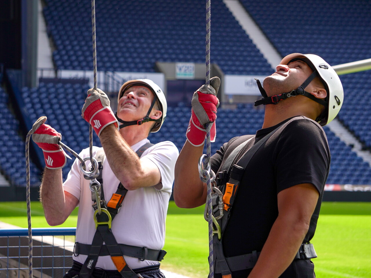 Super Bob Taylor and Nigel Quashie wearing helmets, harnesses and safety gloves prepare for an abseil at The Hawthorns, looking up at the ropes with the stadium seating visible behind them.