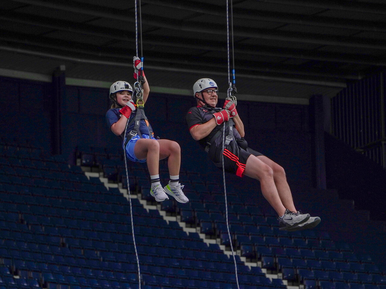 Two participants wearing helmets and safety harnesses descend on ropes inside a sports arena, suspended high above rows of blue stadium seats during a supervised abseiling activity.