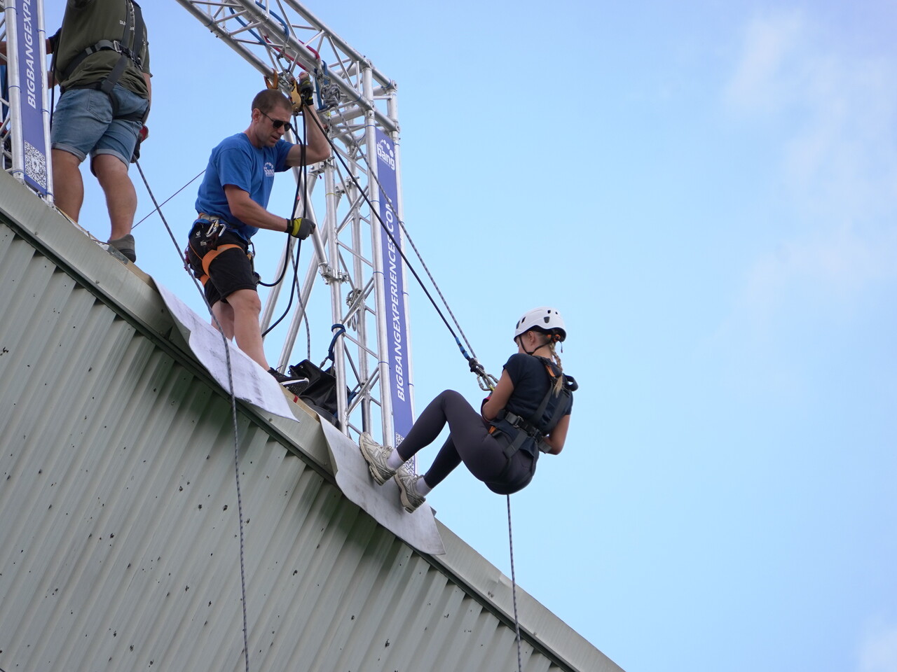 Participant wearing a helmet and safety harness begins an abseil from the roof of a stadium, guided by an instructor standing beside the rope system under a clear blue sky.