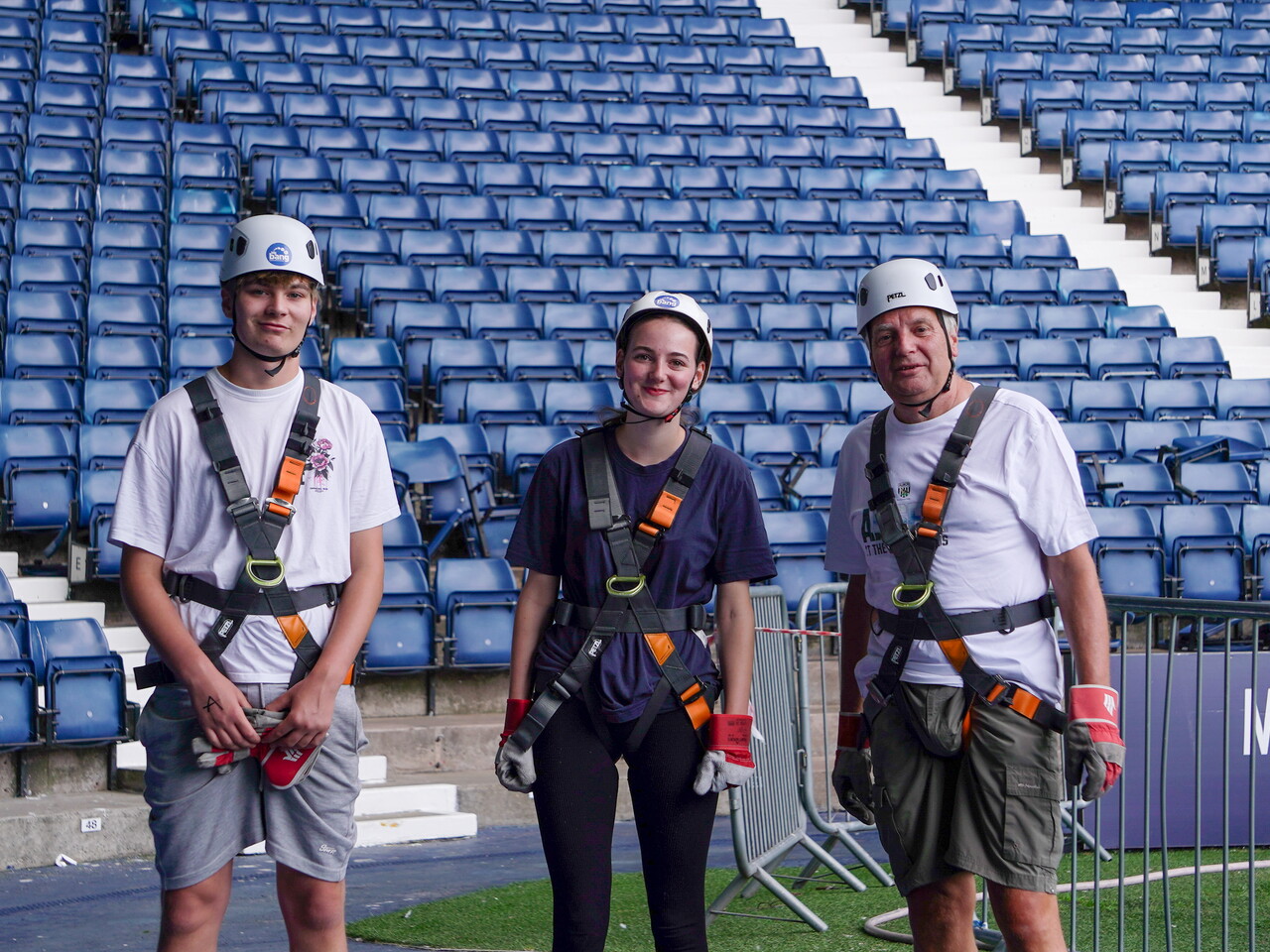 Three participants wearing helmets and safety harnesses pose for a photo on the pitchside area at The Hawthorns after completing an abseil.