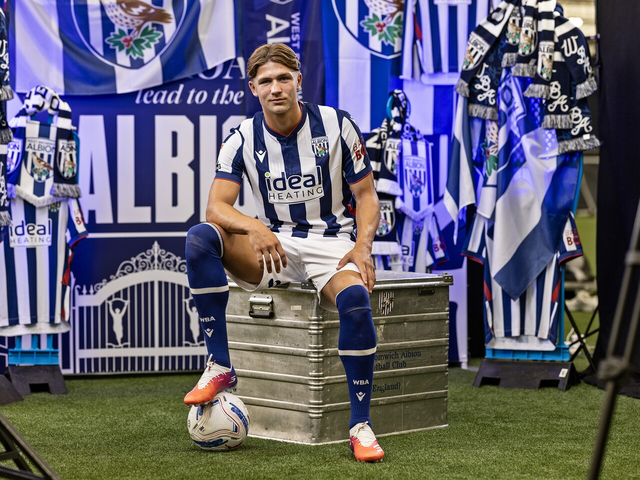 Cole Deeming in the home kit with his foot on a ball while sat on a kit skip in front of an Albion back drop with plenty of WBA branding
