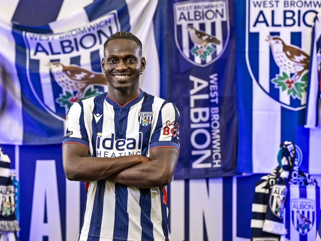 Ousmane Diakité in the home kit smiling at the camera with his arms folded while stood in front of an Albion back drop with plenty of WBA branding