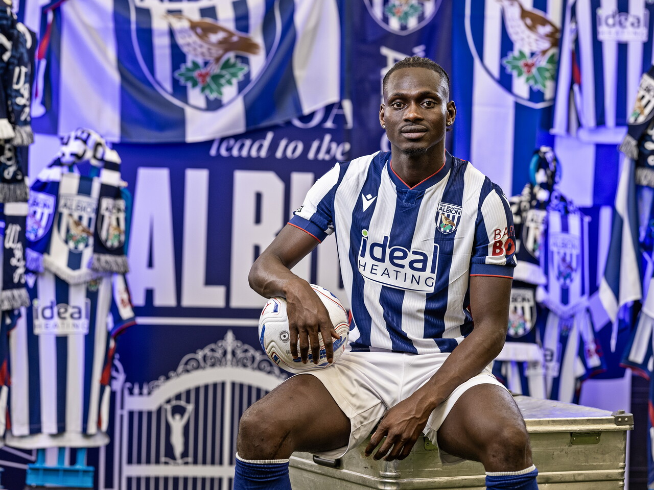 Ousmane Diakité in the home kit holding a ball while sat on a kit skip in front of an Albion back drop with plenty of WBA branding