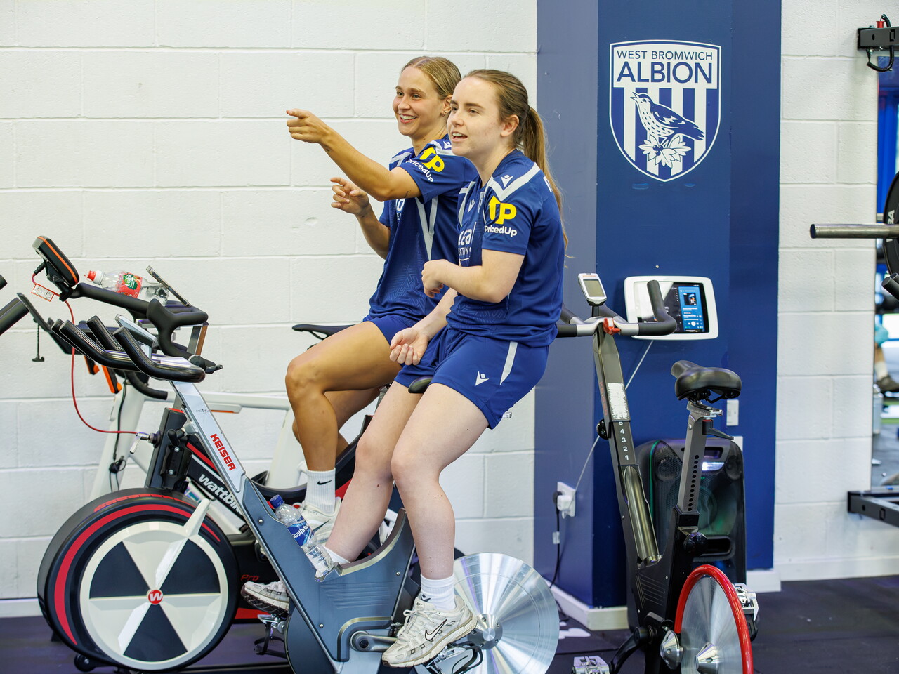 Mia Sorrentino and Ella Haughey sat on a bike in the gym laughing and smiling 