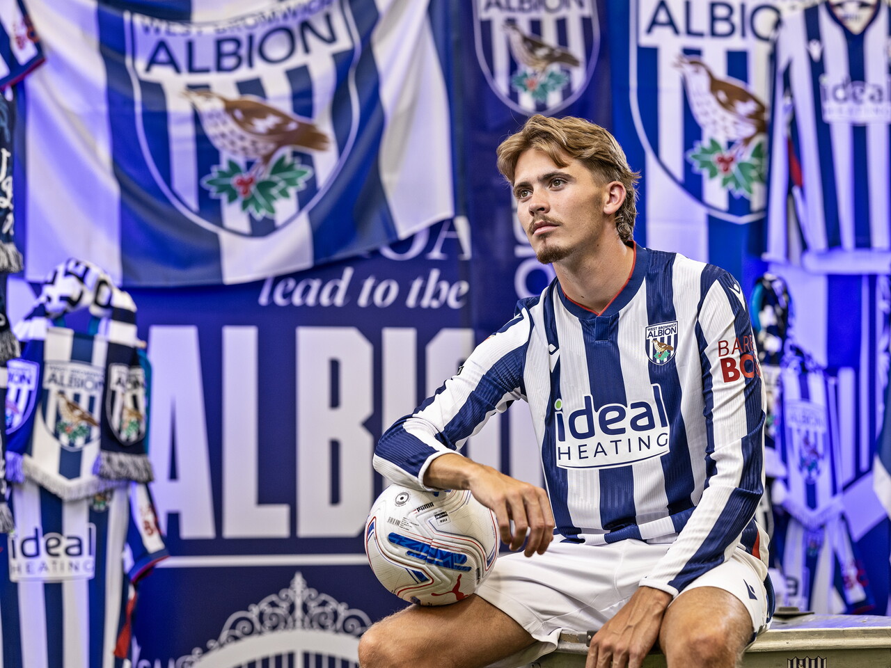 Isaac Price in the home kit holding a ball while sat on a kit skip in front of an Albion back drop with plenty of WBA branding