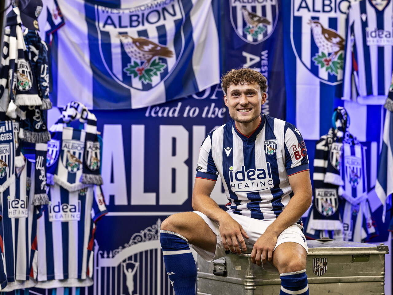 Callum Styles in the home kit smiling at the camera while sat on a kit skip in front of an Albion back drop with plenty of WBA branding