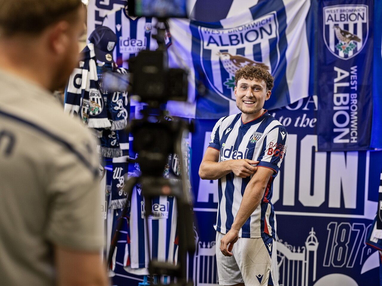 Callum Styles in the home kit smiling at the camera while stood in front of an Albion back drop with plenty of WBA branding