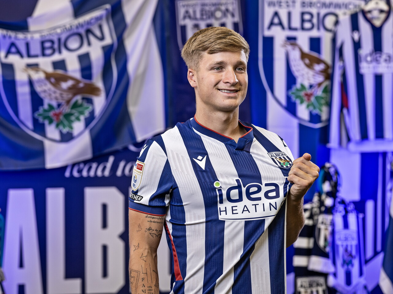 JJ Morgan in the home kit smiling at the camera while stood in front of an Albion back drop with plenty of WBA branding