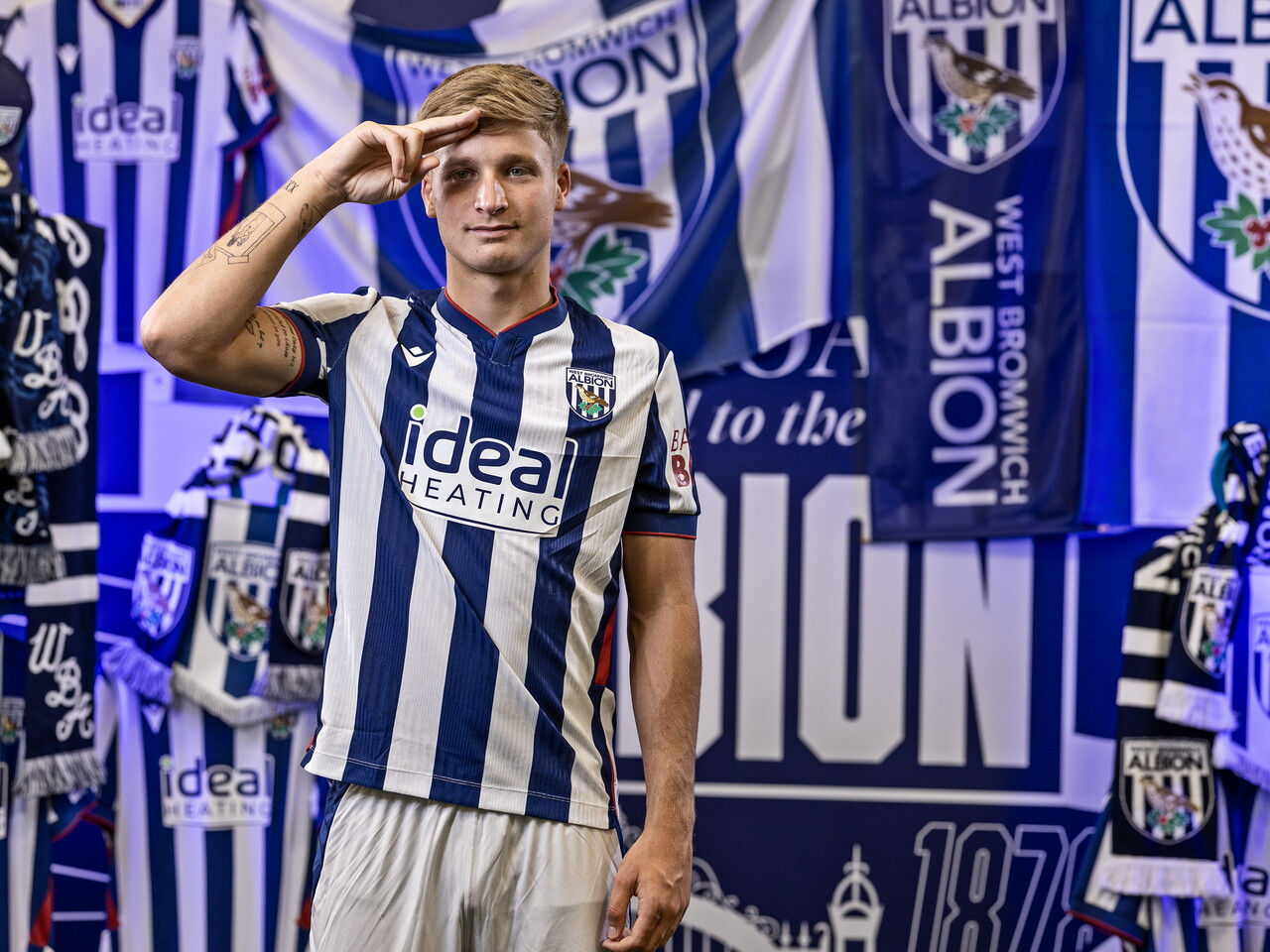 JJ Morgan in the home kit saluting towards the camera while stood in front of an Albion back drop with plenty of WBA branding