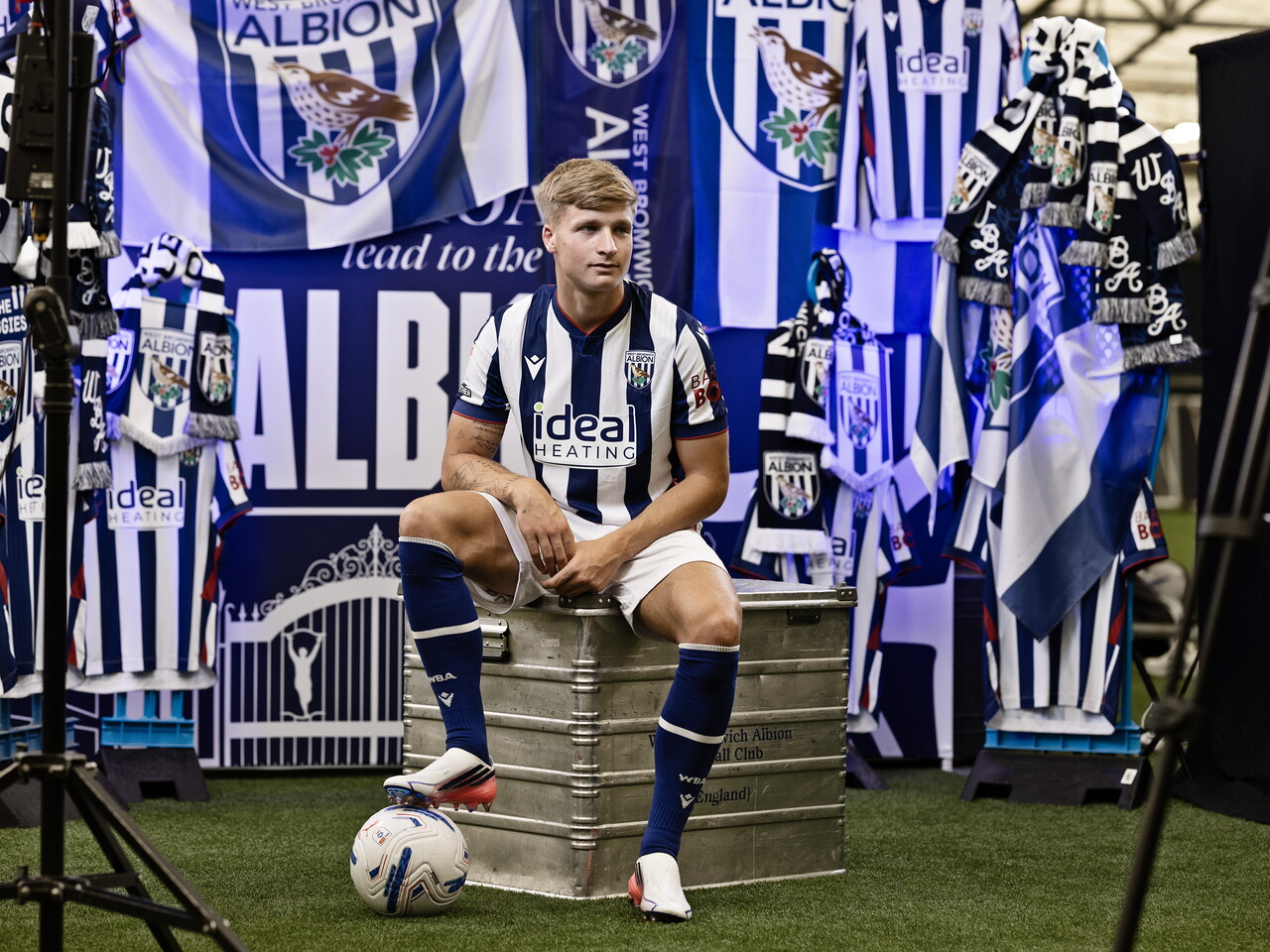 JJ Morgan in the home kit with his foot on a ball while sat on a kit skip in front of an Albion back drop with plenty of WBA branding
