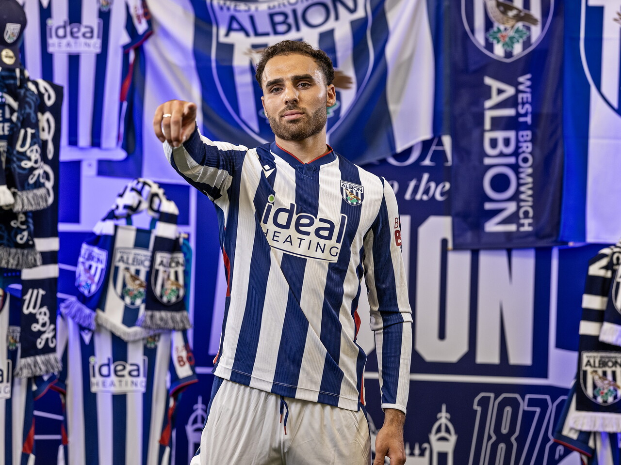 Tammer Bany in the home kit pointing at the camera while stood in front of an Albion back drop with plenty of WBA branding
