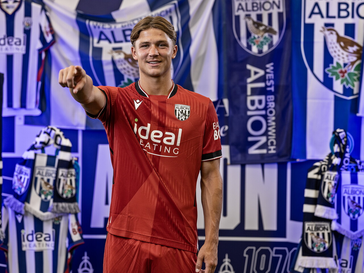 Cole Deeming in the red away kit pointing at the camera while stood in front of an Albion back drop with plenty of WBA branding