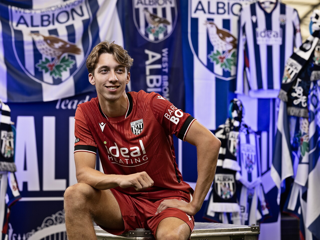 Harry Whitwell in the red away kit smiling at the camera while sat on a kit skip in front of an Albion back drop with plenty of WBA branding