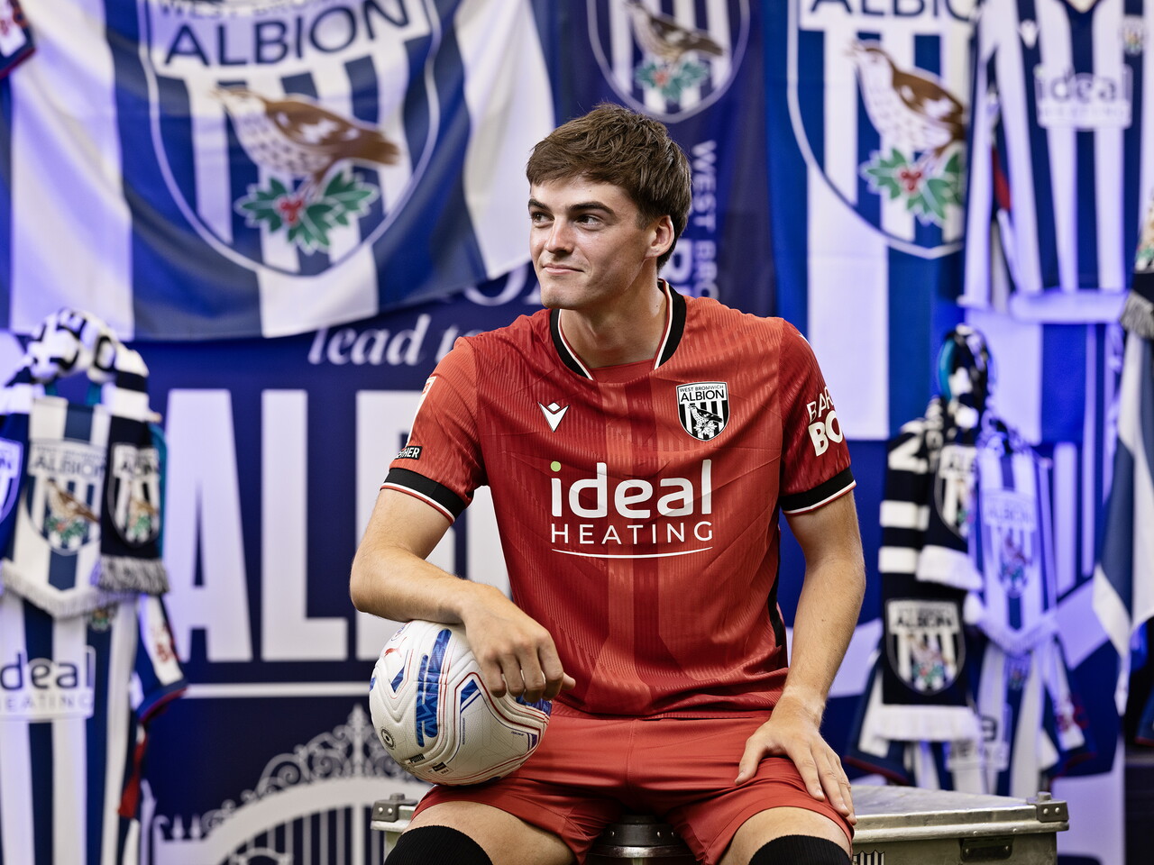 Alex Williams in the red away kit holding a ball and looking away from camera while sat in front of an Albion back drop with plenty of WBA branding