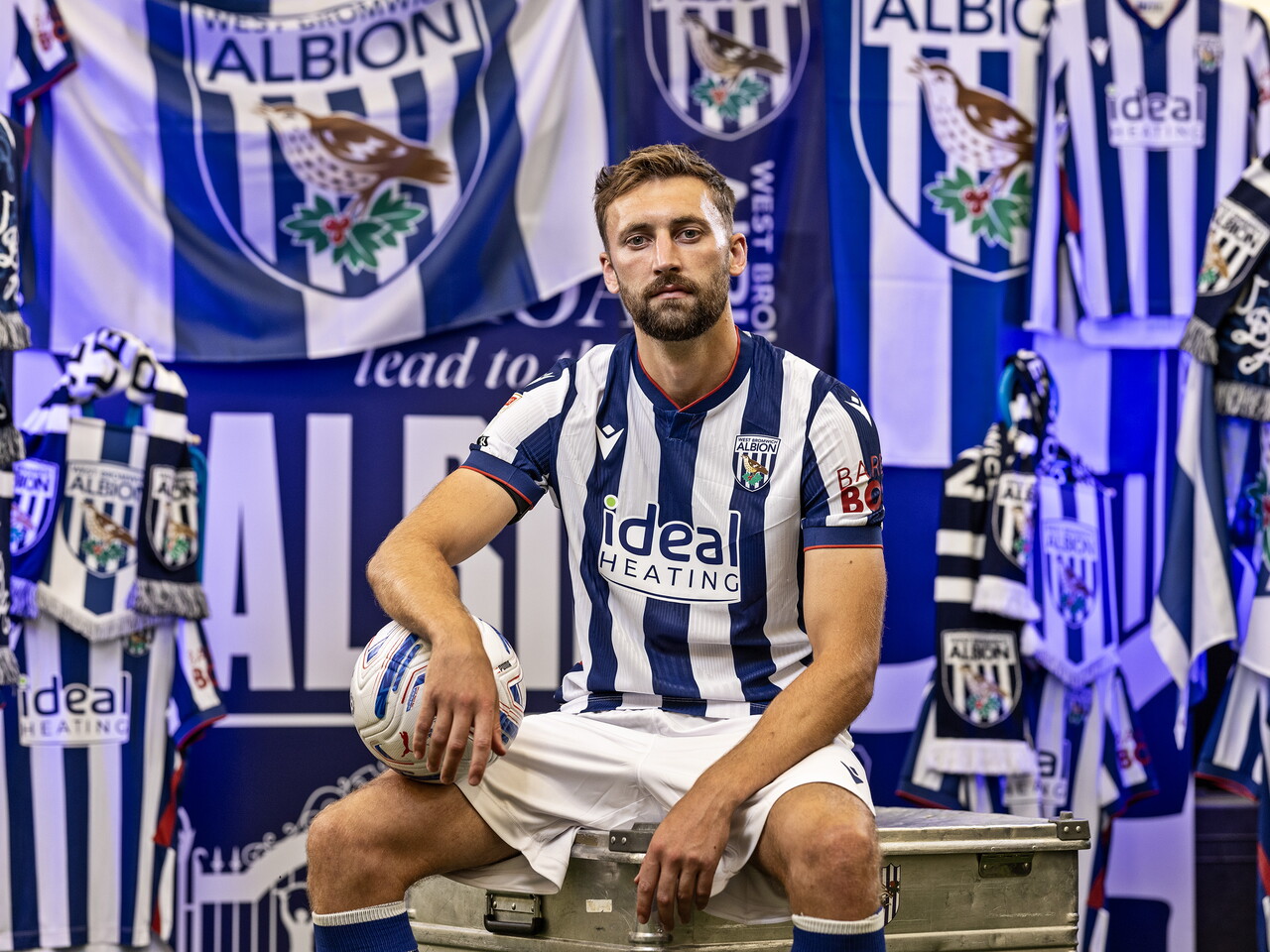 Nat Phillips in the home kit holding a ball and looking at the camera while sat in front of an Albion back drop with plenty of WBA branding