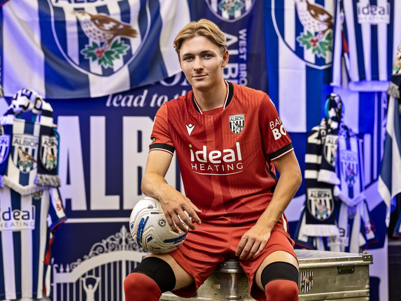 Ollie Bostock in the red away kit smiling at the camera and holding a ball while sat in front of an Albion back drop with plenty of WBA branding