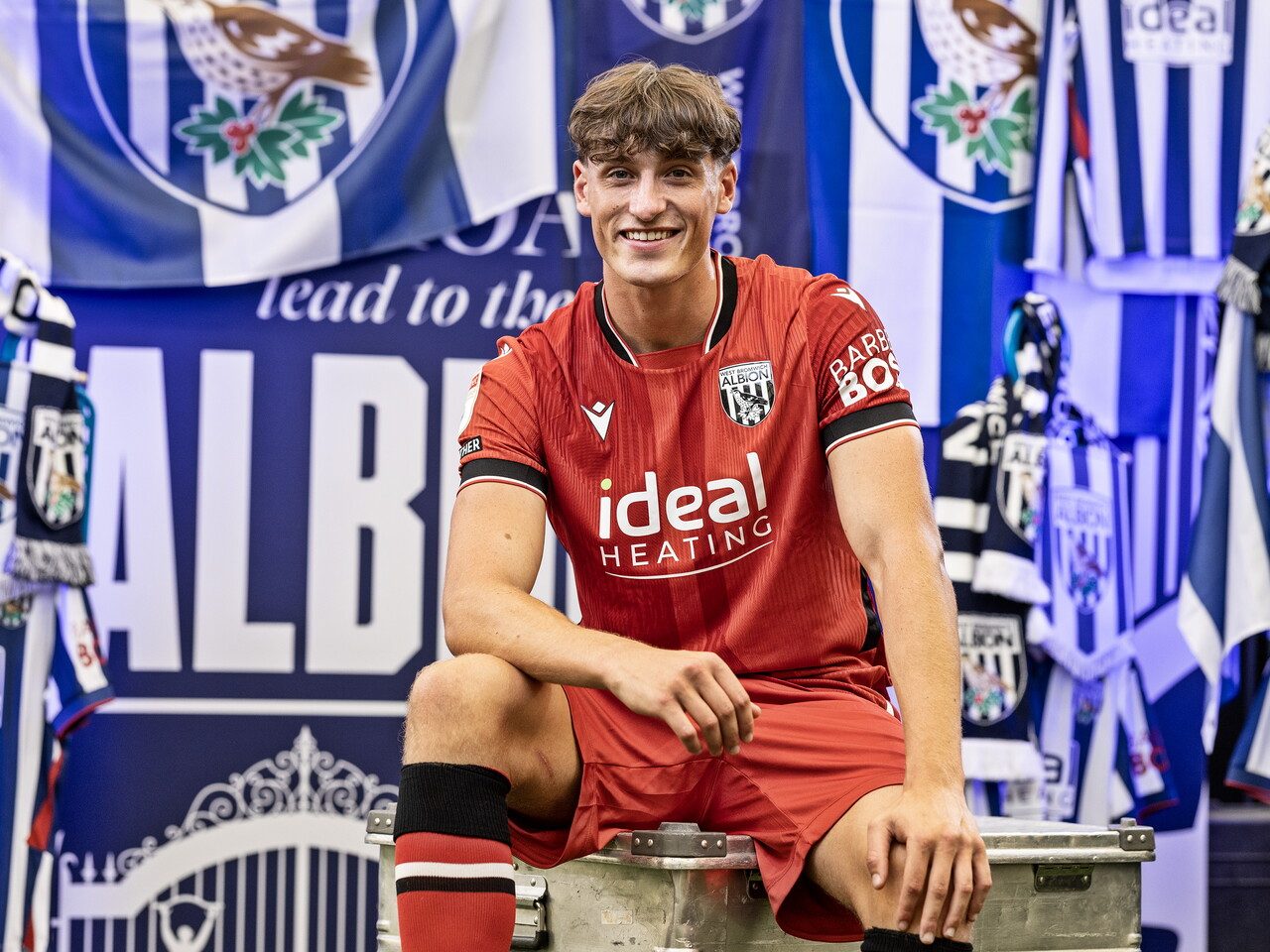 Barney Stewart in the red away kit smiling at the camera while sat in front of an Albion back drop with plenty of WBA branding