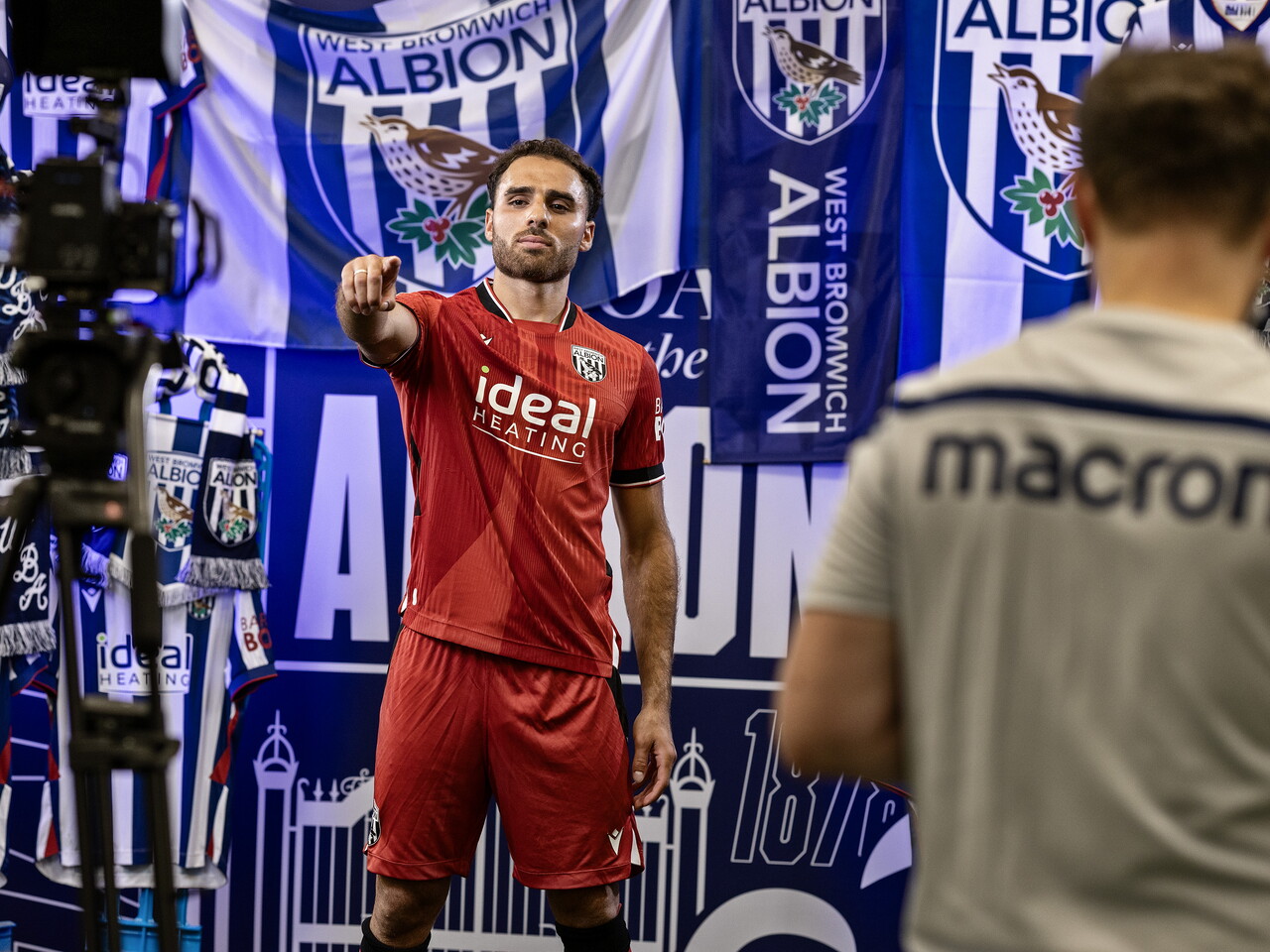 Tammer Bany in the red away kit pointing towards the camera while stood in front of an Albion back drop with plenty of WBA branding
