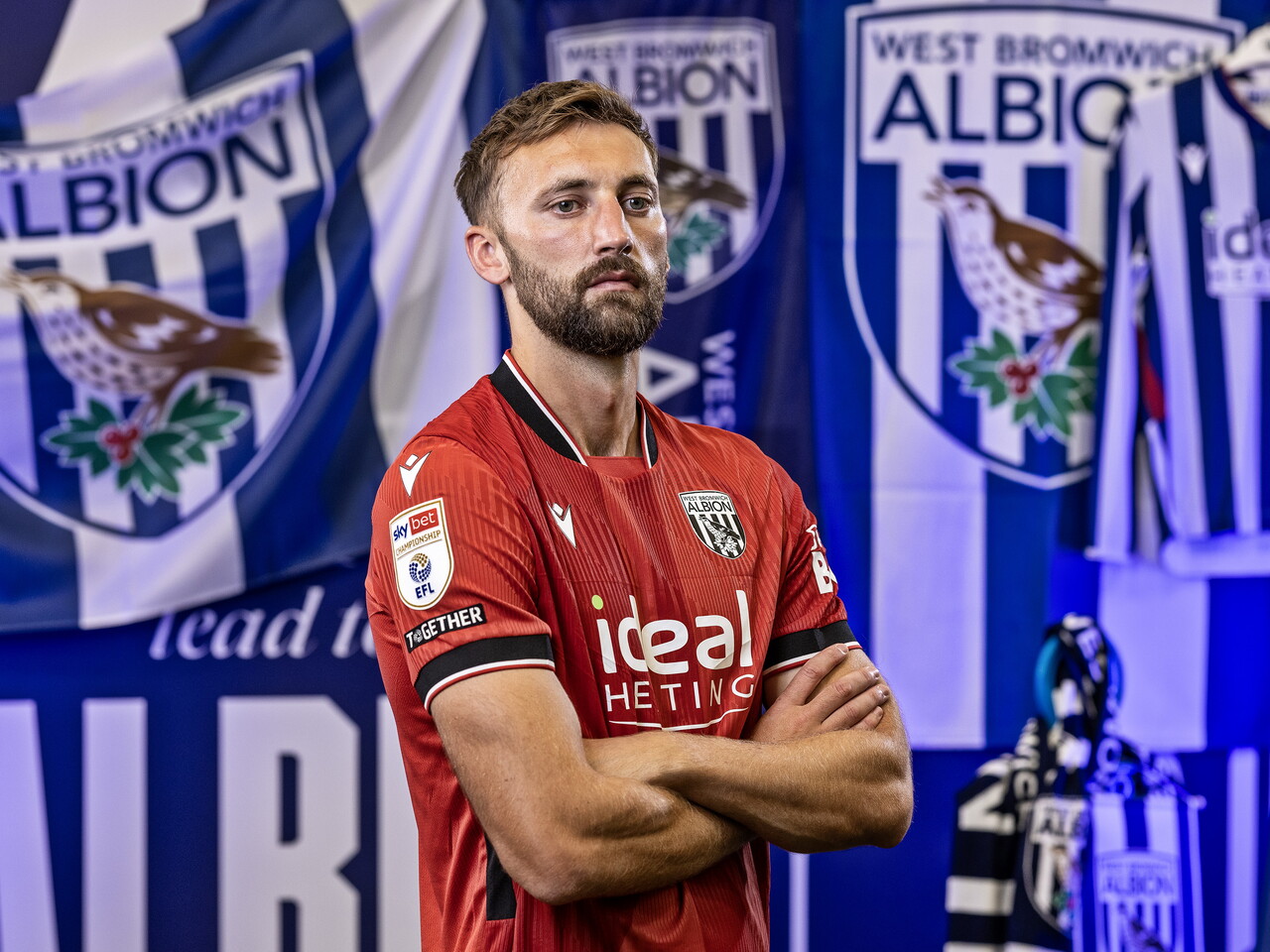 Nat Phillips in the red away kit looking at the camera with his arms folded while stood in front of an Albion back drop with plenty of WBA branding