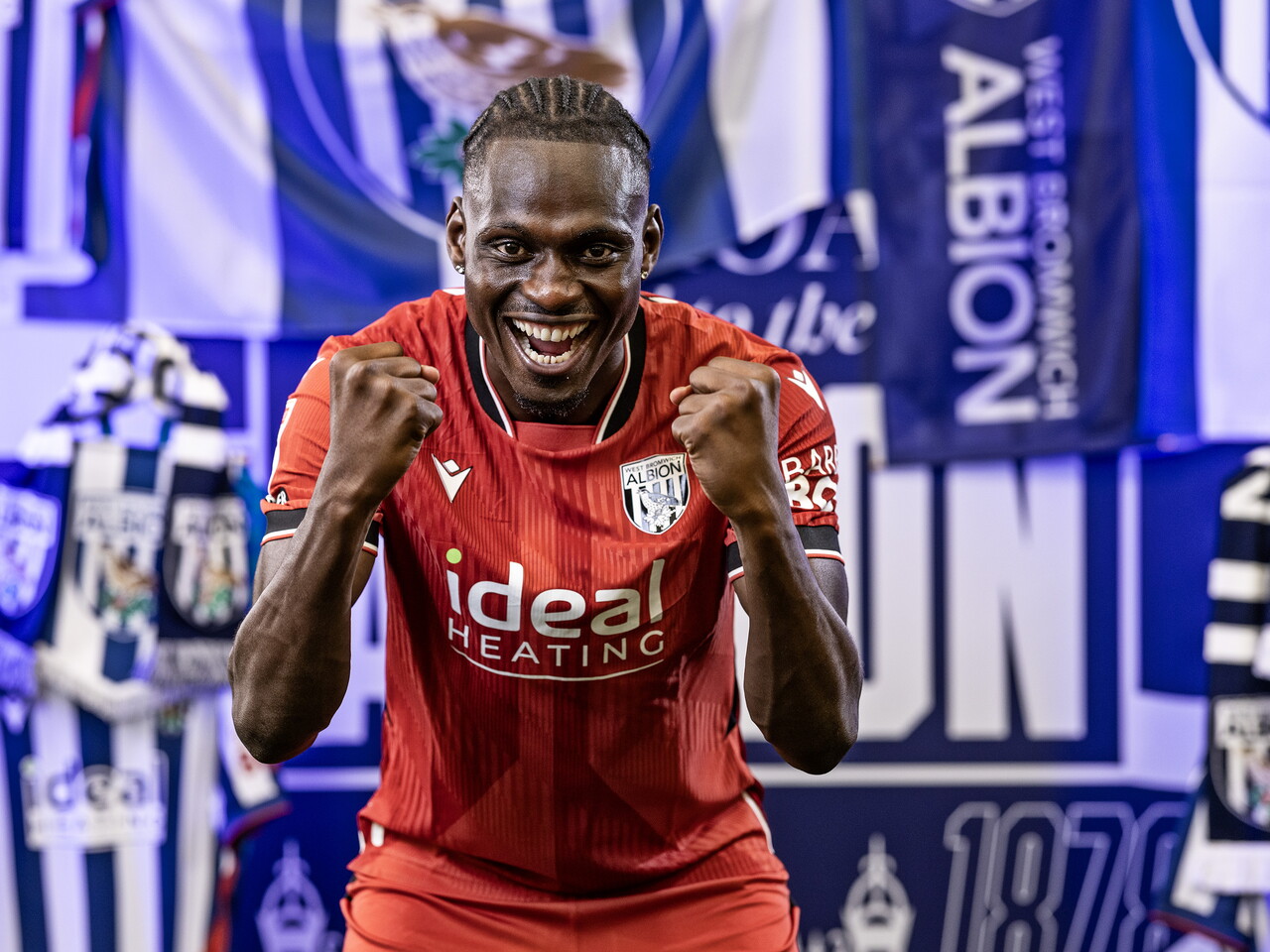 Ousmane Diakité in the red away kit celebrating towards the camera while stood in front of an Albion back drop with plenty of WBA branding