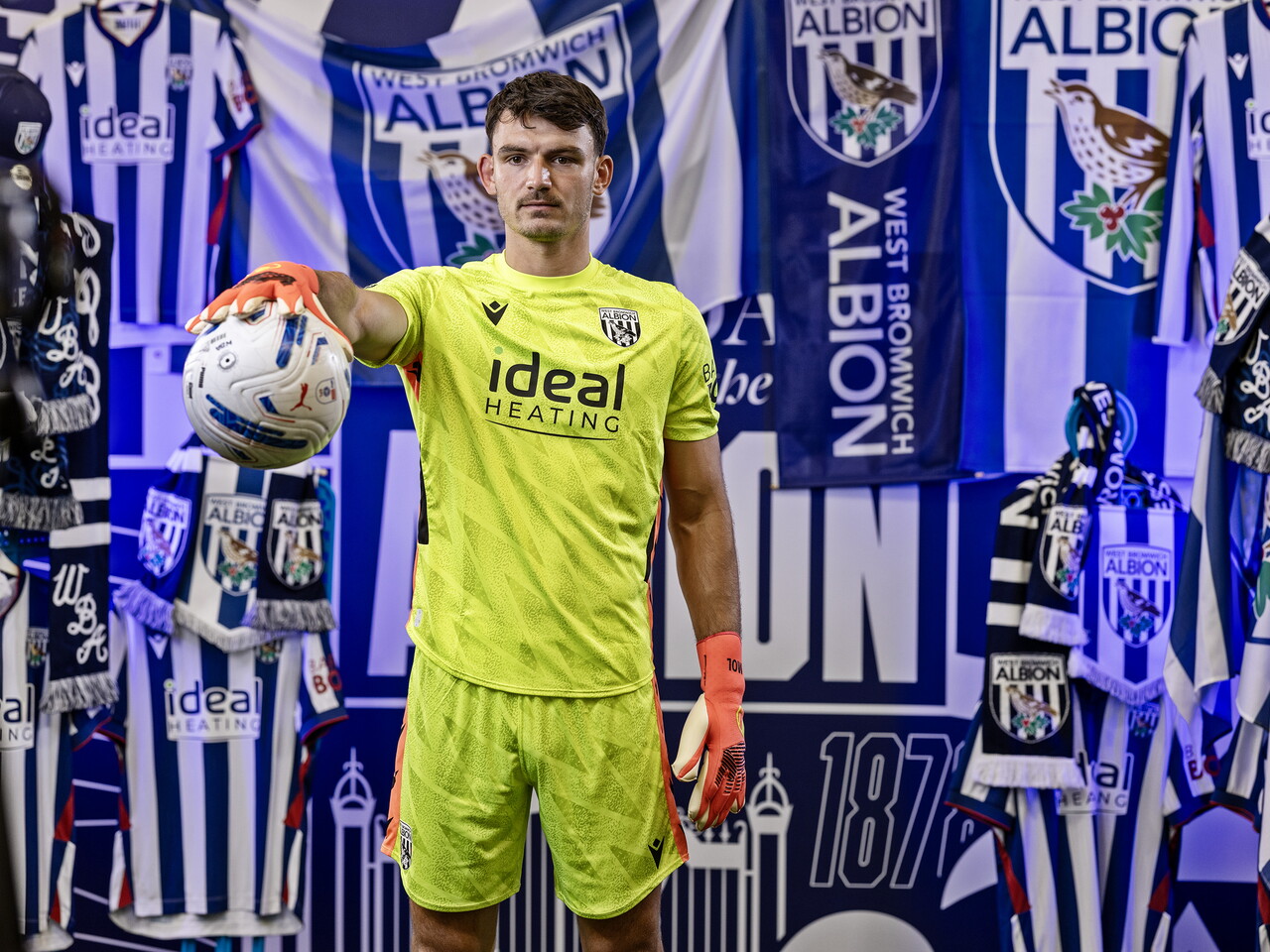 Max O'Leary in the yellow GK kit looking at the camera and holding a ball while stood in front of an Albion back drop with plenty of WBA branding