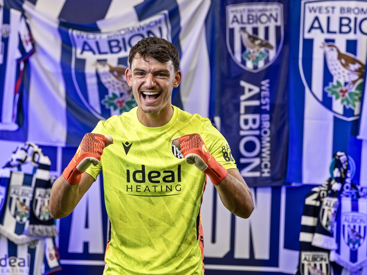 Max O'Leary in the yellow GK kit celebrating towards the camera while stood in front of an Albion back drop with plenty of WBA branding