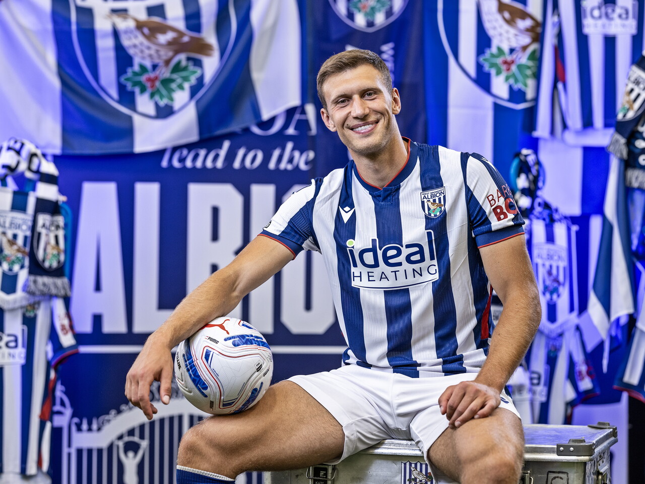 Krystian Bielik sat smiling at the camera while holding a ball in front of an Albion back drop with plenty of WBA branding