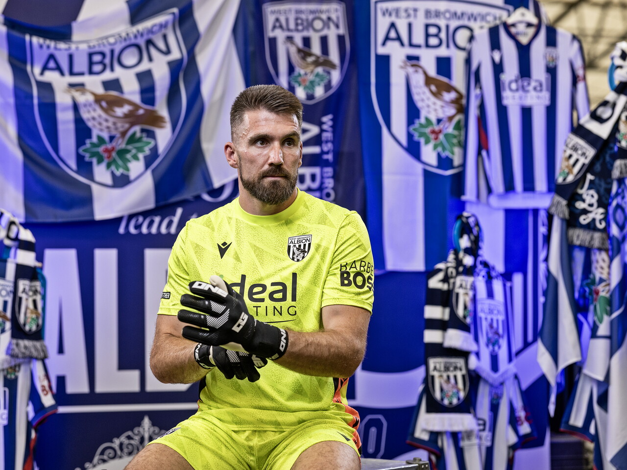 Matt Ingram in the yellow GK kit looking away from camera while sat in front of an Albion back drop with plenty of WBA branding