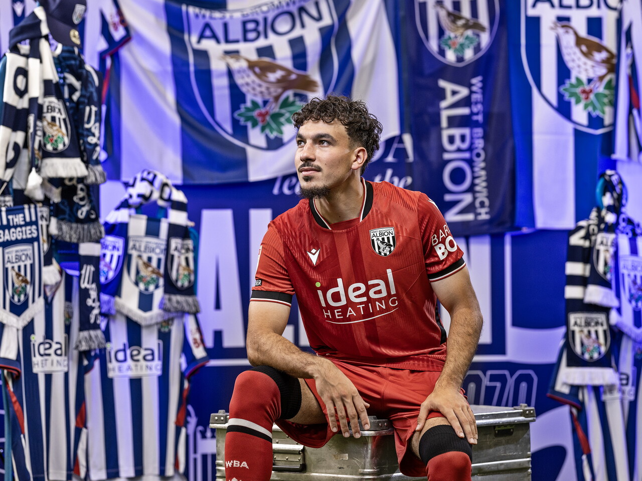 Nolan Galves in the red away kit looking away from camera while sat in front of an Albion back drop with plenty of WBA branding