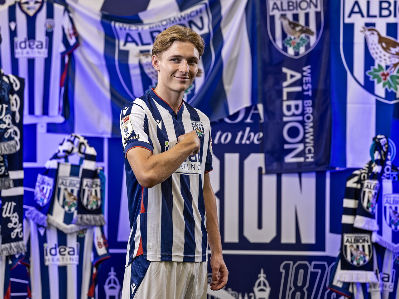 Ollie Bostock in the home kit smiling at the camera while stood in front of an Albion back drop with plenty of WBA branding