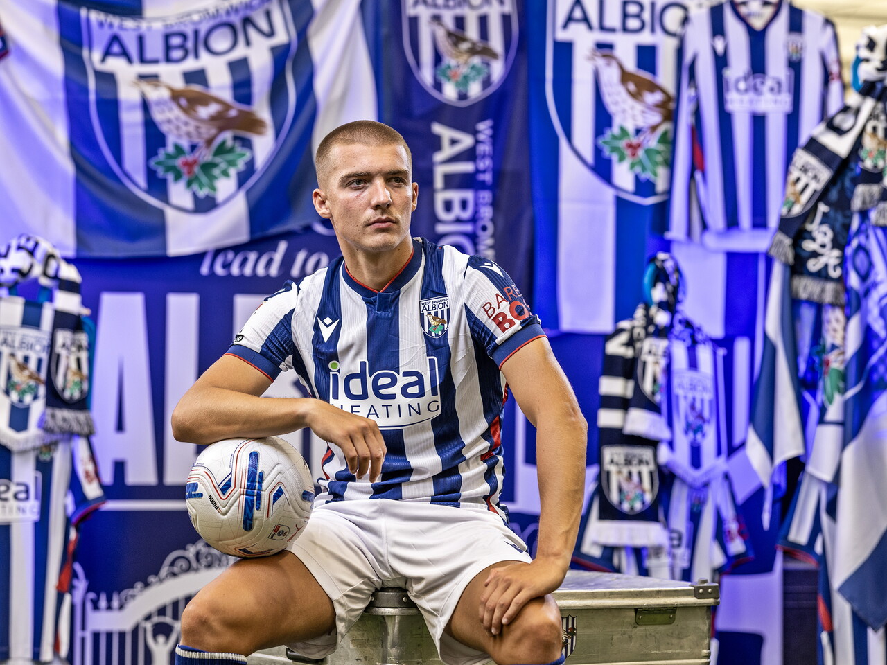 Alfie Gilchrist in the home kit holding a ball while sat in front of an Albion back drop with plenty of WBA branding