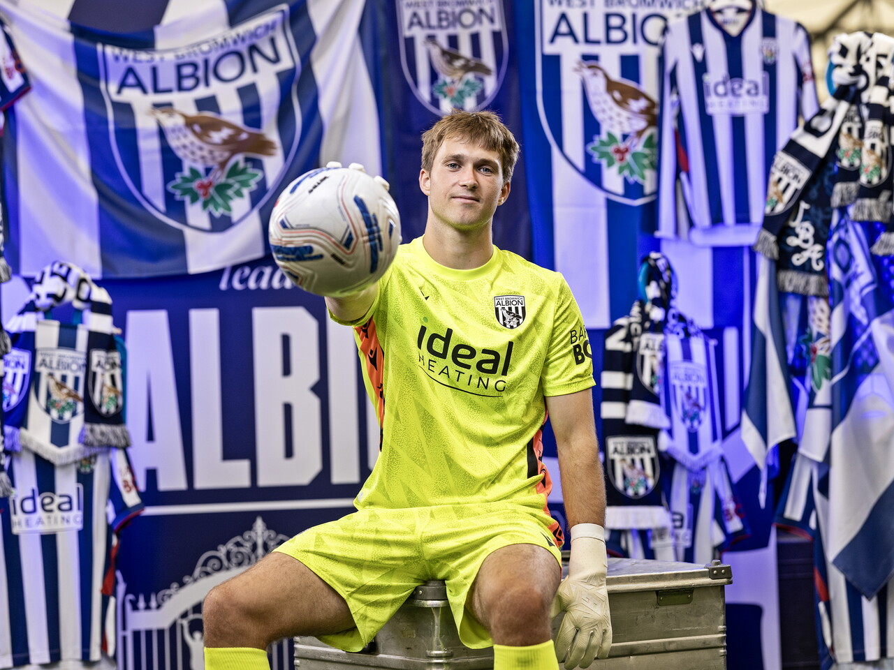 Joe Wallis in the yellow GK kit smiling at the camera and holding a ball while sat in front of an Albion back drop with plenty of WBA branding