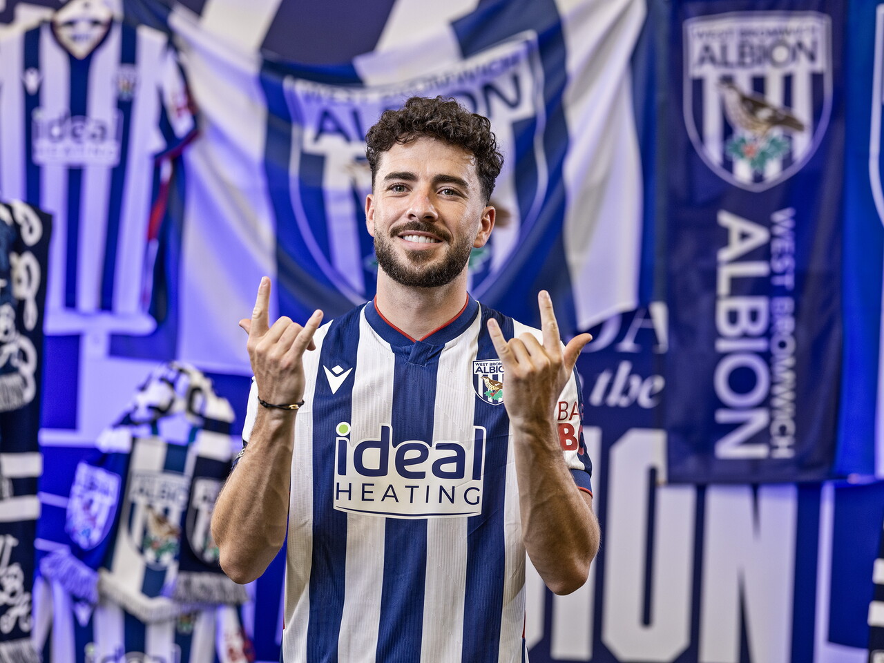 Mikey Johnston in the home kit smiling at the camera while stood in front of an Albion back drop with plenty of WBA branding