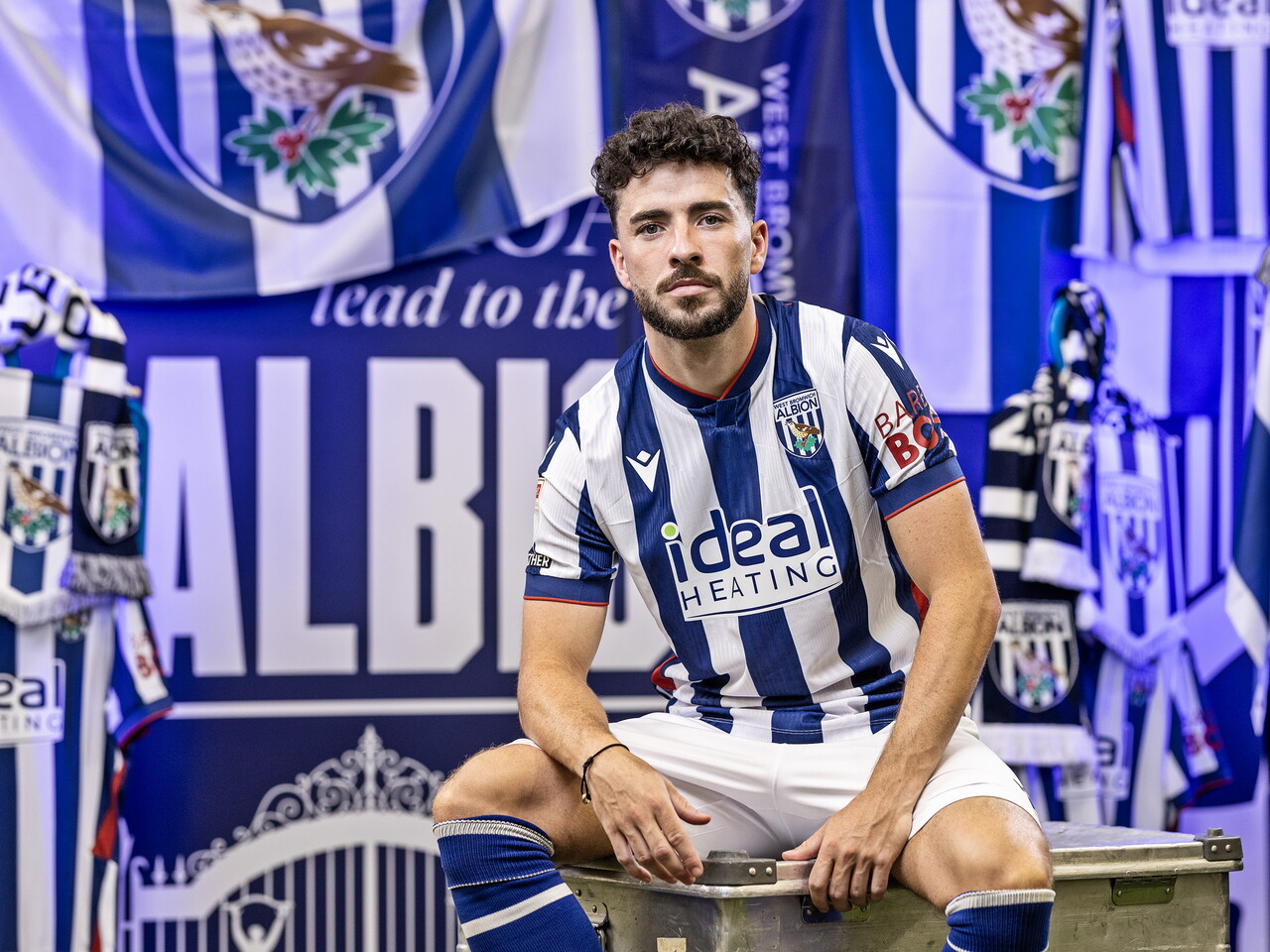 Mikey Johnston in the home kit looking at the camera while sat in front of an Albion back drop with plenty of WBA branding