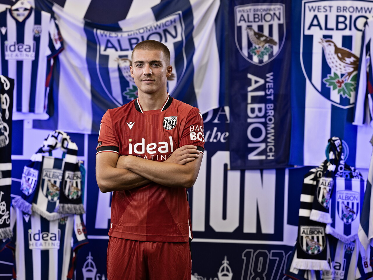 Alfie Gilchrist in the red away kit looking at the camera with his arms folded while stood in front of an Albion back drop with plenty of WBA branding