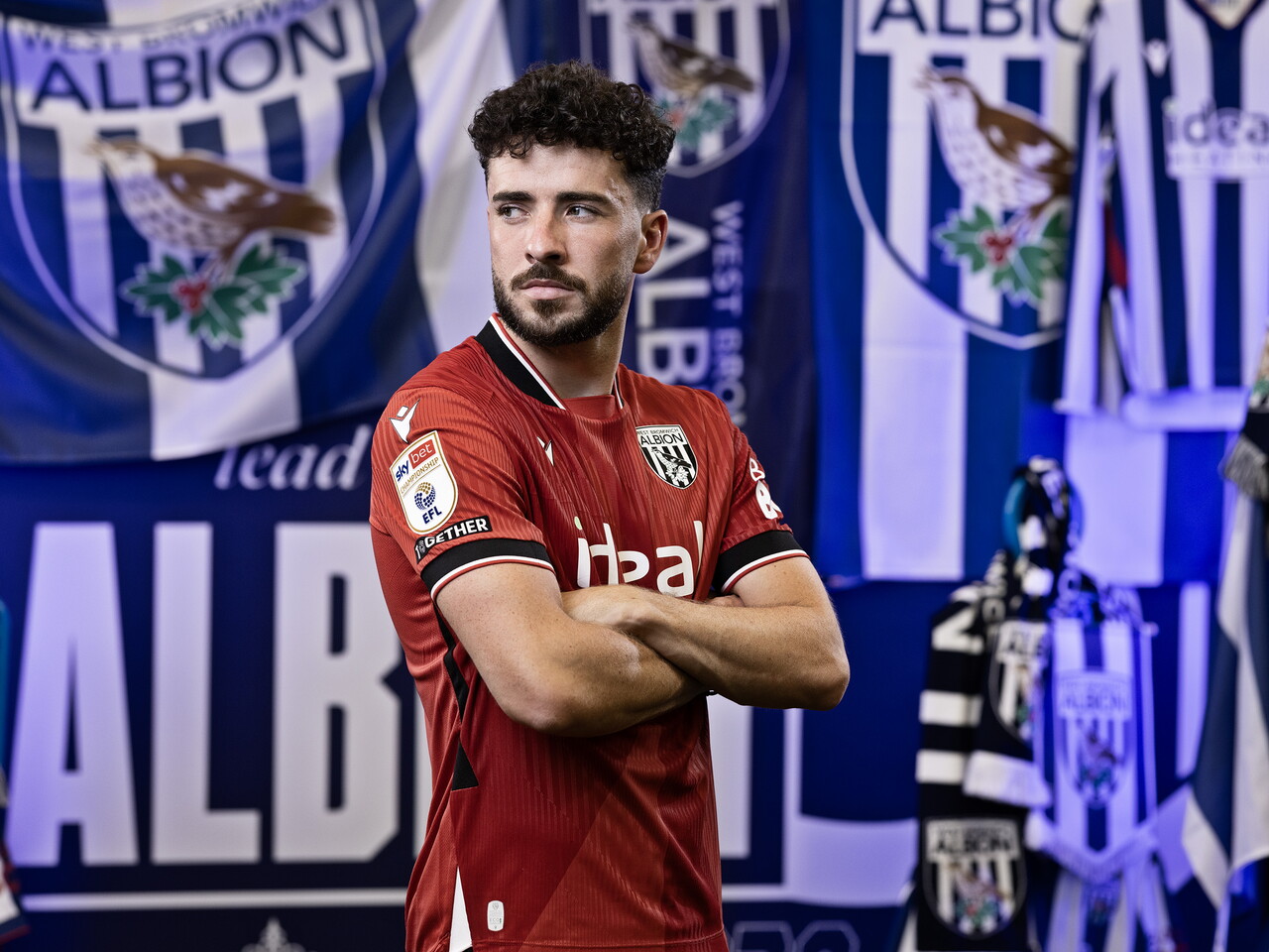 Mikey Johnston in the red away kit looking away from camera with his arms folded while stood in front of an Albion back drop with plenty of WBA branding