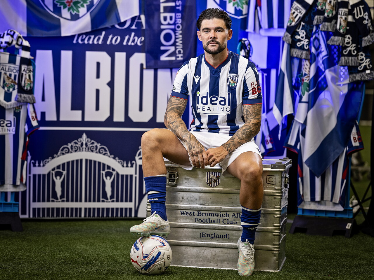 Alex Mowatt sat on a kit skip with his foot on a ball in front of an Albion back drop with plenty of WBA branding